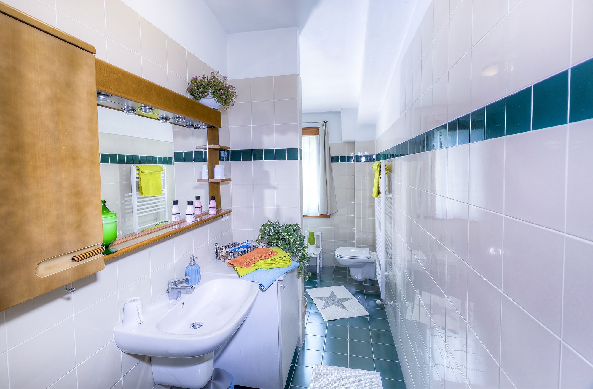 Un baño con lavamanos, espejo e inodoro. Hay repisas con artículos de aseo y una planta.