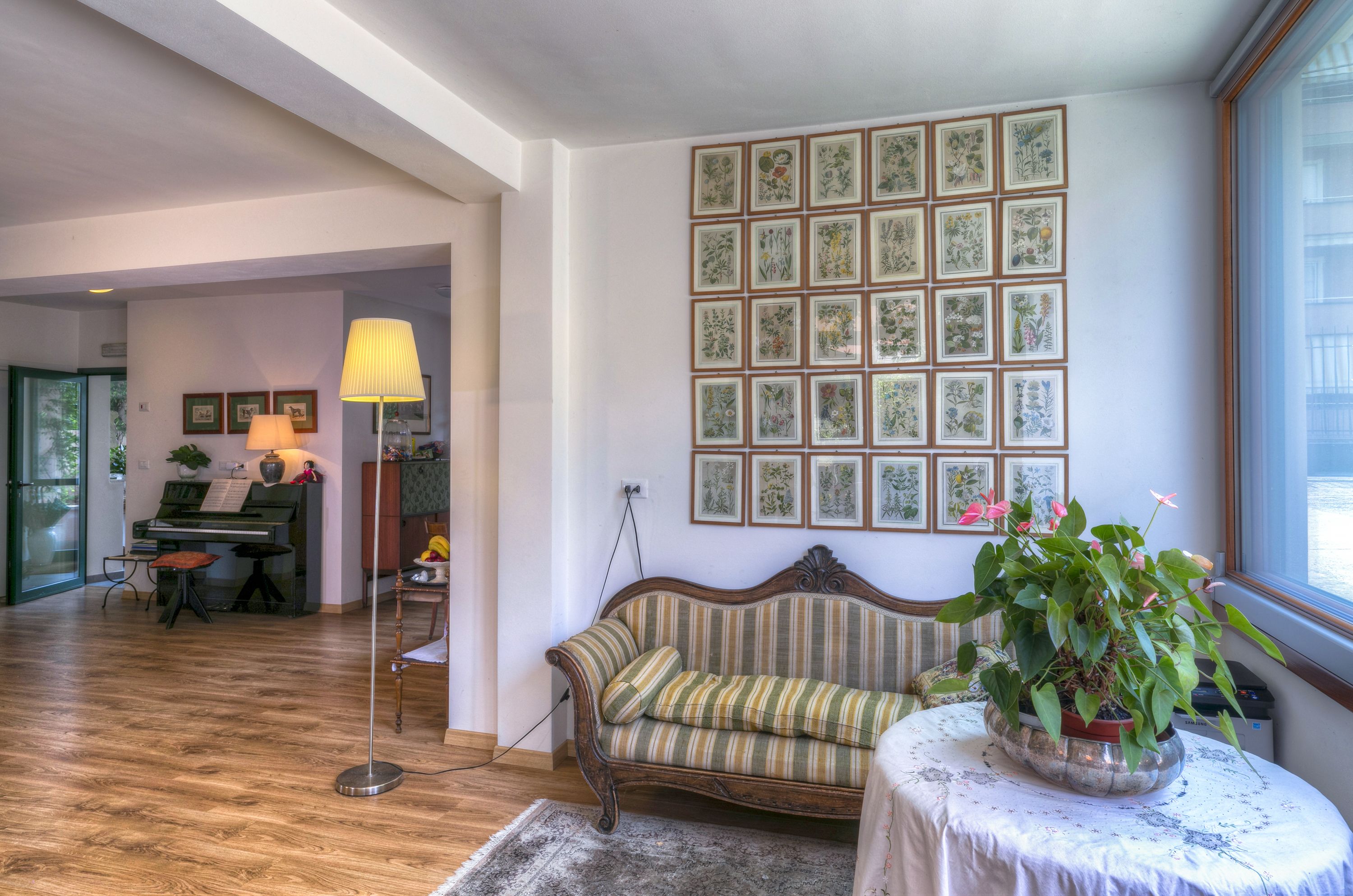 Ein Wohnzimmer mit einem Klavier, einer Couch, einer Lampe und einer Wand mit gerahmten Pflanzenbildern.