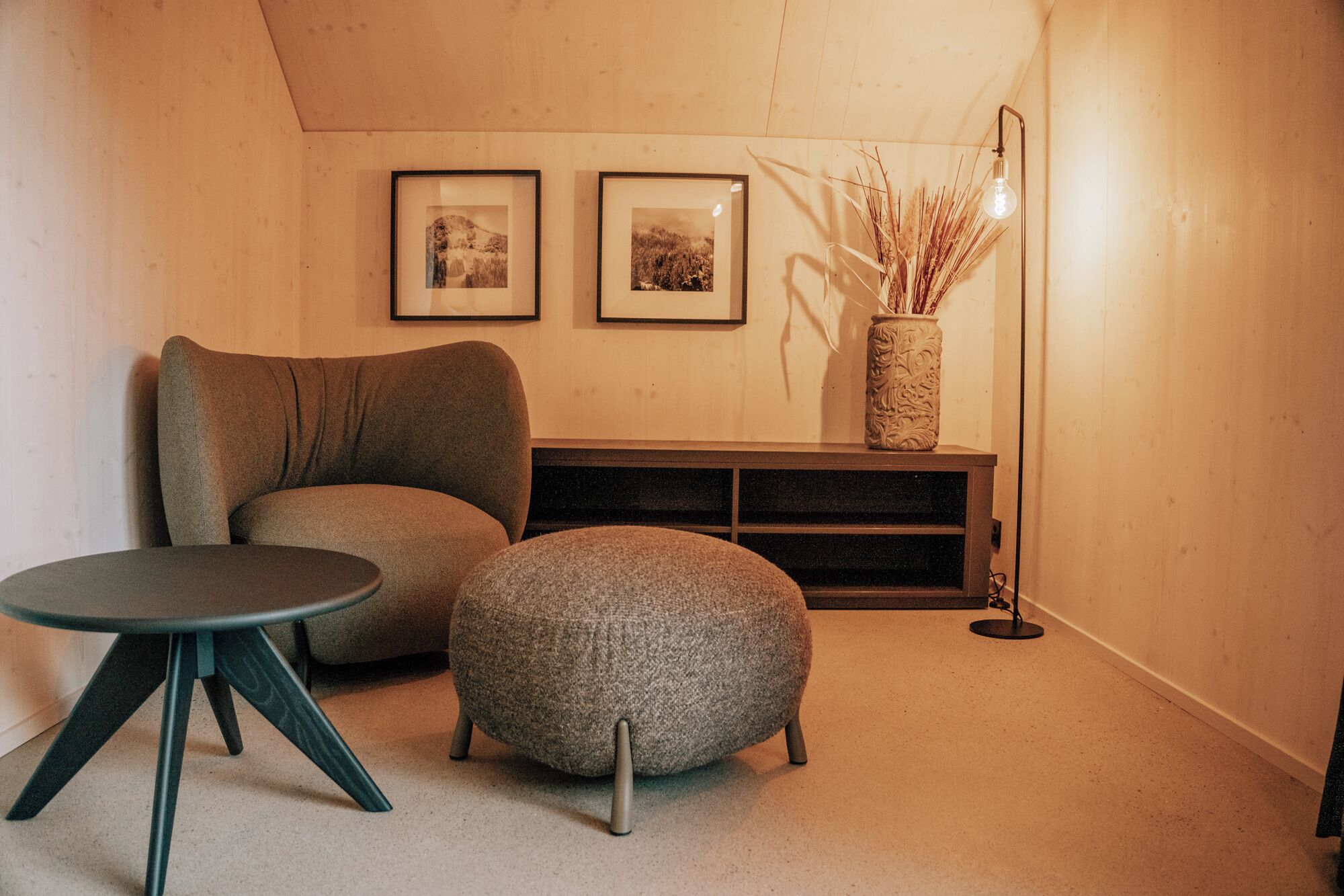 Une pièce chaleureuse avec un canapé, un pouf et une petite table. Deux tableaux encadrés ornent le mur, et un vase contenant des plantes séchées est posé sur une console.