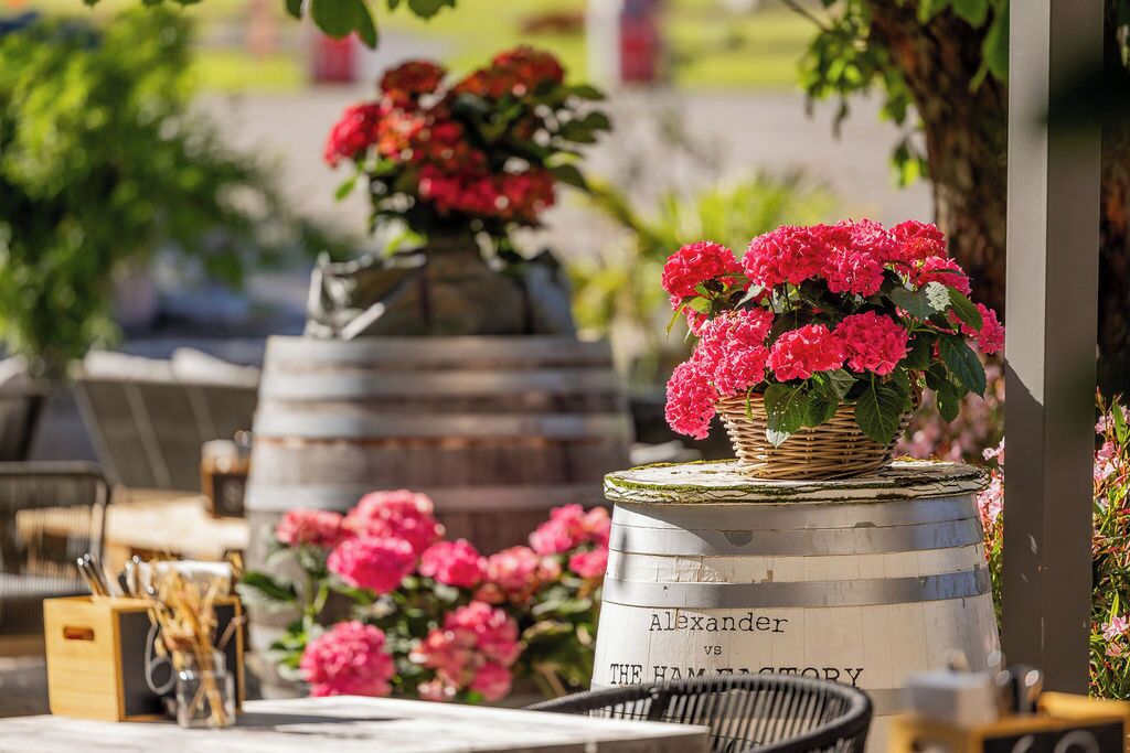 A dining area with a table set for two, a wicker basket with flowers, and a barrel labeled 'Alexander vs The Hamptons.'