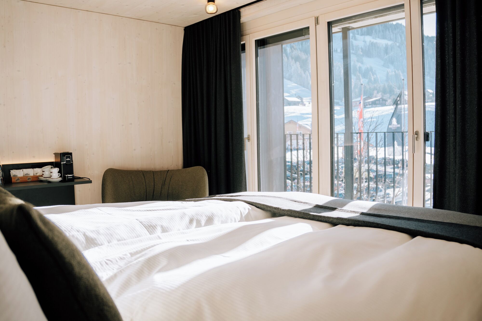 Ein Hotelzimmer mit Bett, Stuhl, kleinem Tisch mit Kaffeemaschine und Fenster mit Blick auf eine verschneite Landschaft.