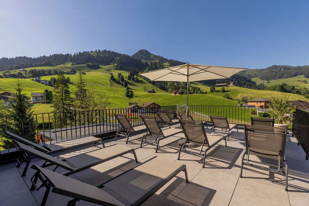 A terrace with sun loungers and a large umbrella, overlooking a green valley and mountains.
