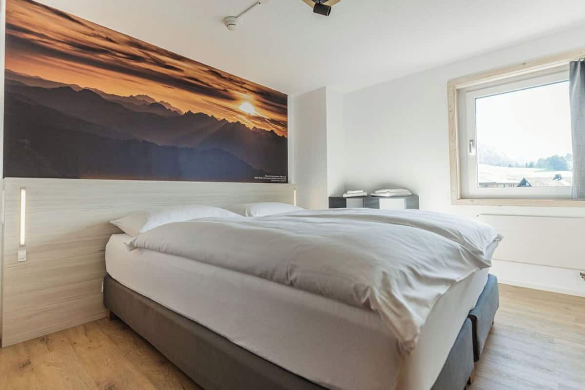 Ein Schlafzimmer mit Bett, Nachttisch, Fenster mit Blick und einem Wandbild.