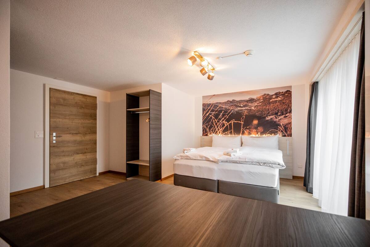 Ein modernes Hotelzimmer mit einem großen Bett, Holzboden, einem Kleiderschrank und einem Gemälde einer Berglandschaft.