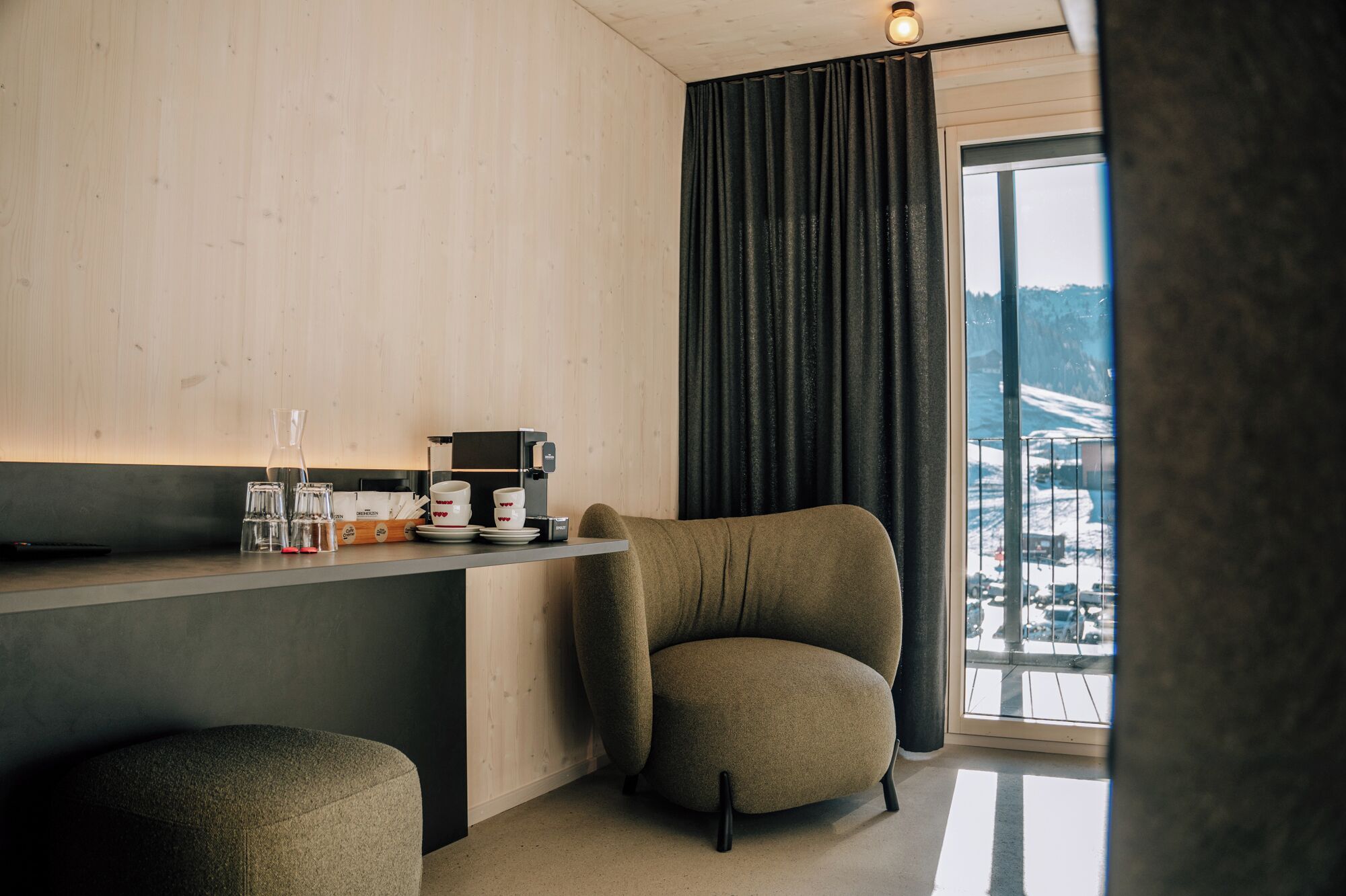 Een moderne kamer met een houten wand, een donker gordijn, een koffieapparaat en uitzicht op een besneeerd landschap door een groot raam.