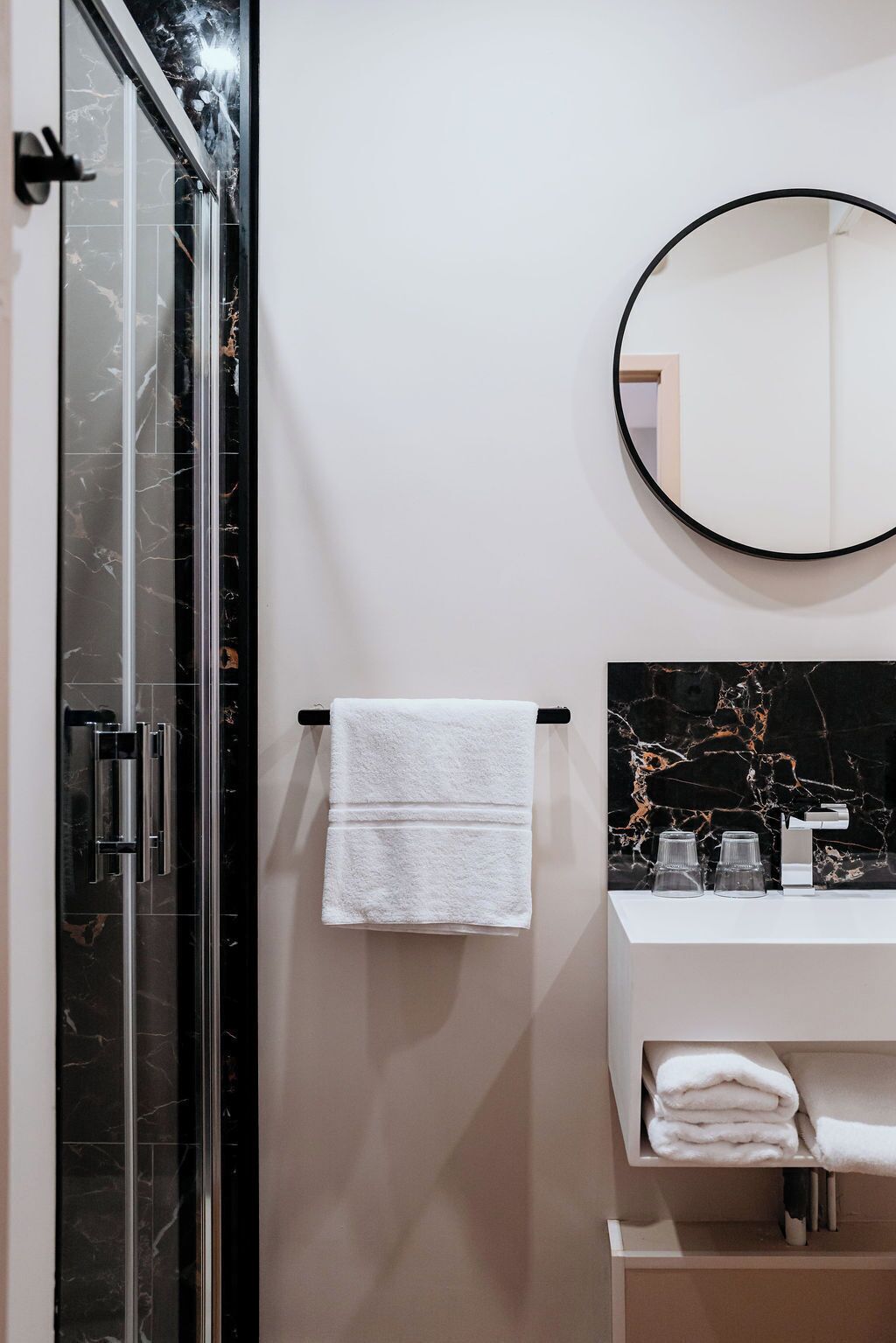 Une salle de bain moderne avec une douche à parois de verre, une serviette blanche suspendue à une porte-serviettes noire, un miroir rond et un plan de toilette en marbre agrémenté de quelques objets en verre.