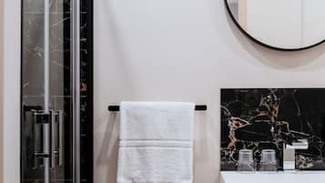 Une salle de bain moderne avec une douche à parois de verre, une serviette blanche suspendue à une porte-serviettes noire, un miroir rond et un plan de toilette en marbre agrémenté de quelques objets en verre.