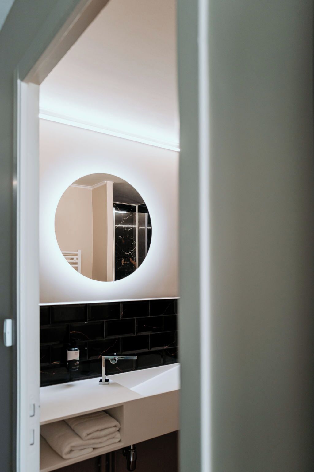 Une salle de bain moderne avec un grand miroir rond, des murs blancs et un pare-feu en carreaux noirs.