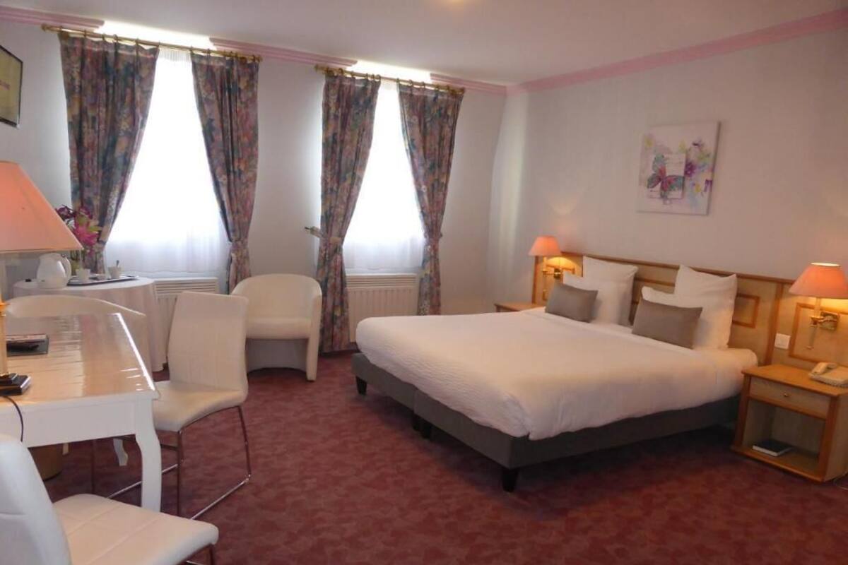 A hotel room with a bed, two bedside tables with lamps, a desk with a chair, and a radiator.