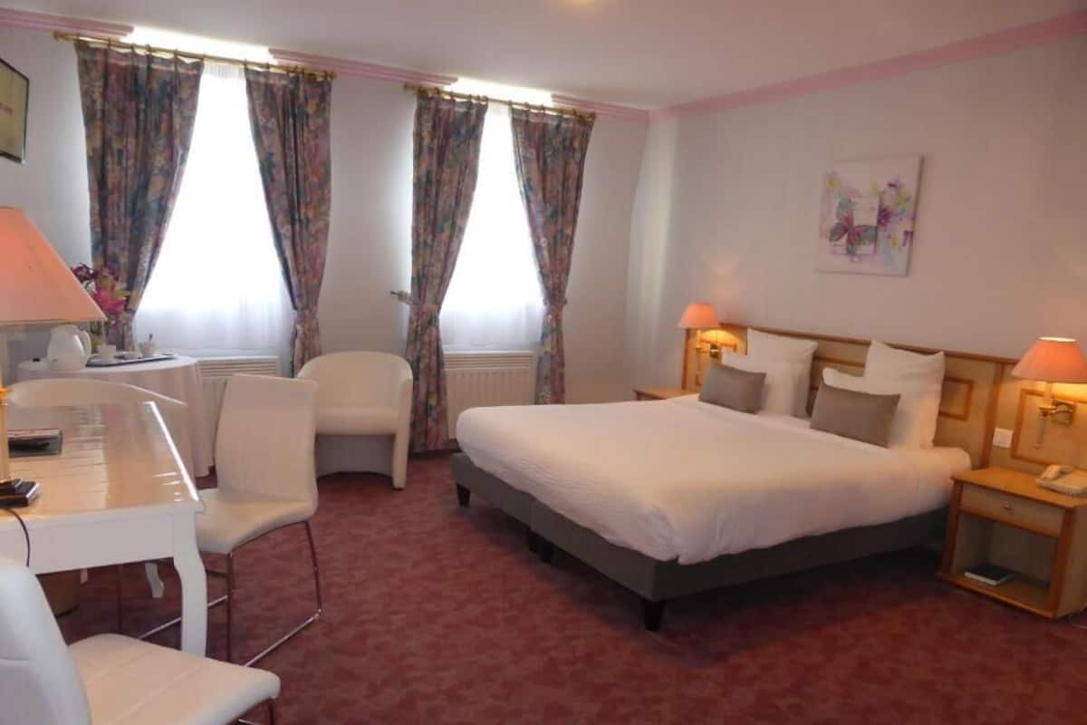 A hotel room with a bed, two bedside tables with lamps, a desk with a chair, and a radiator.