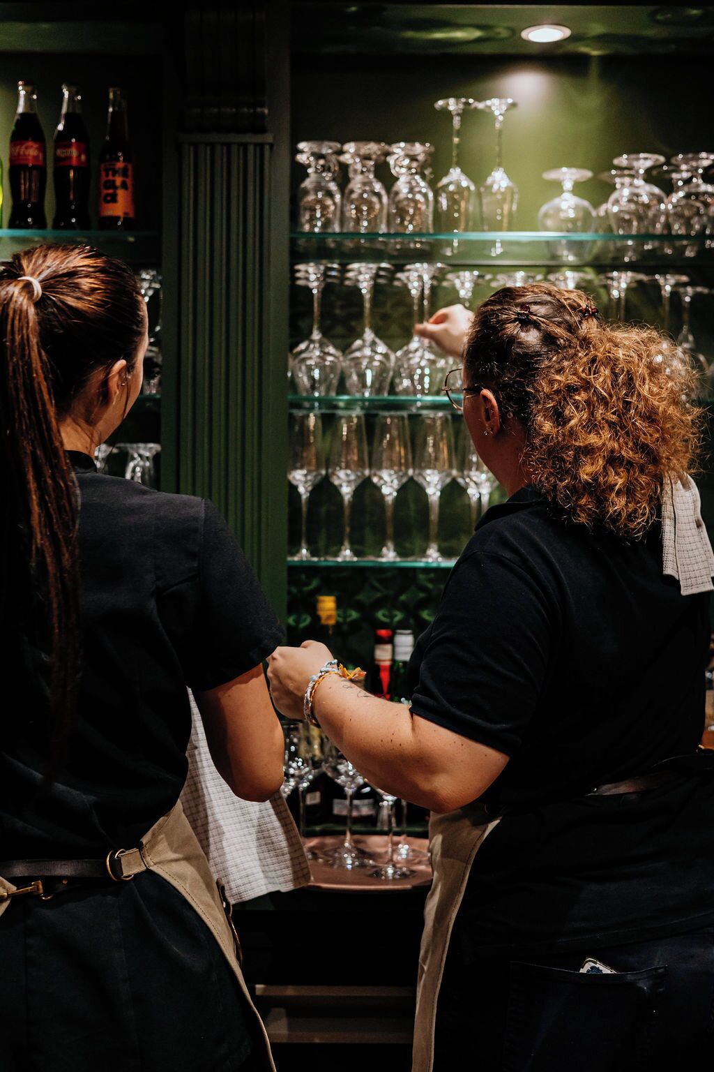 Two individuals in a bar setting, one arranging glasses on a shelf and the other reaching for a bottle.