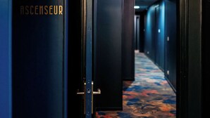 Un pasillo con piso alfombrado y una puerta que indica 'ASCENSEUR'.