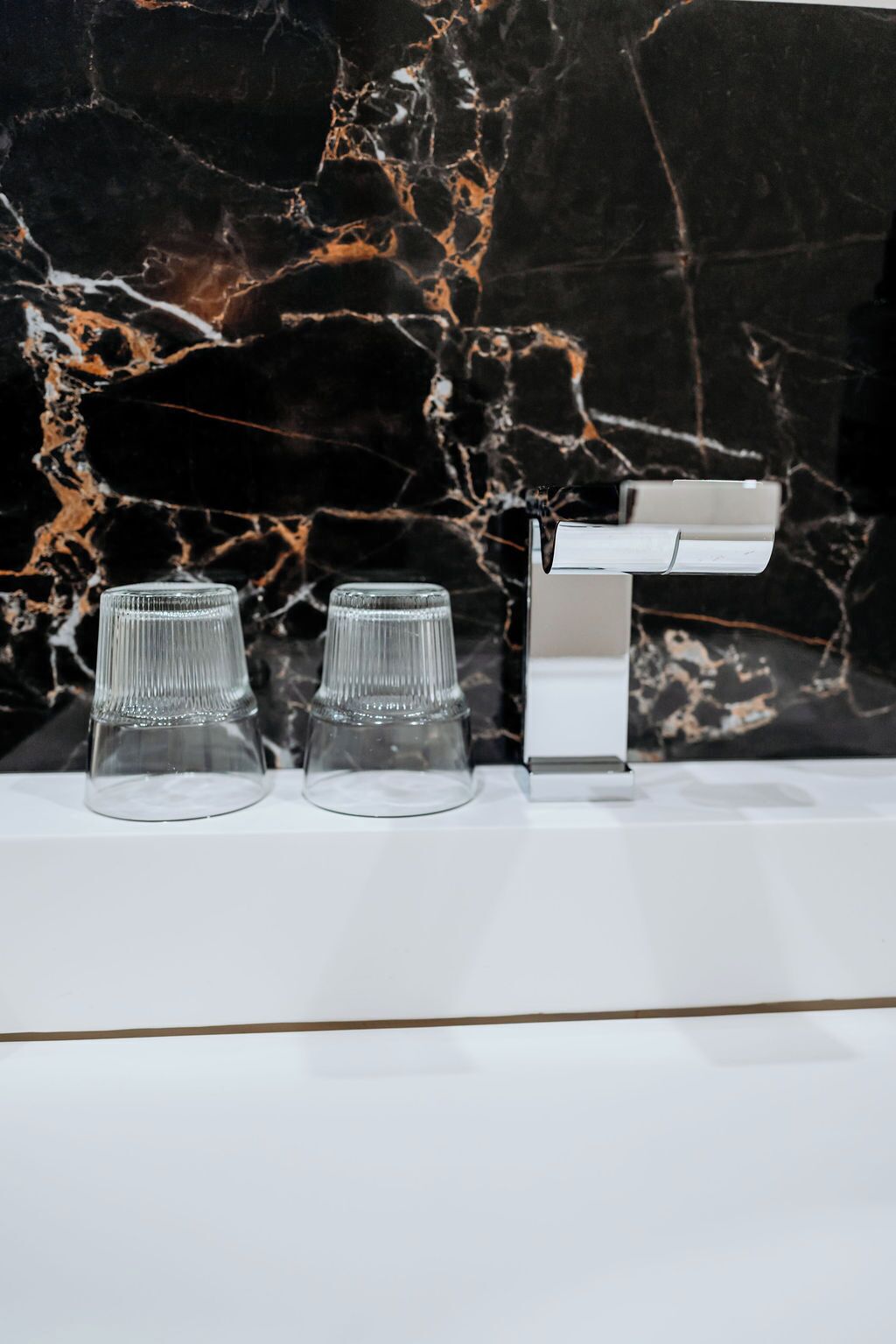 A bathroom with a marble backsplash, a white sink, and two clear glass cups.
