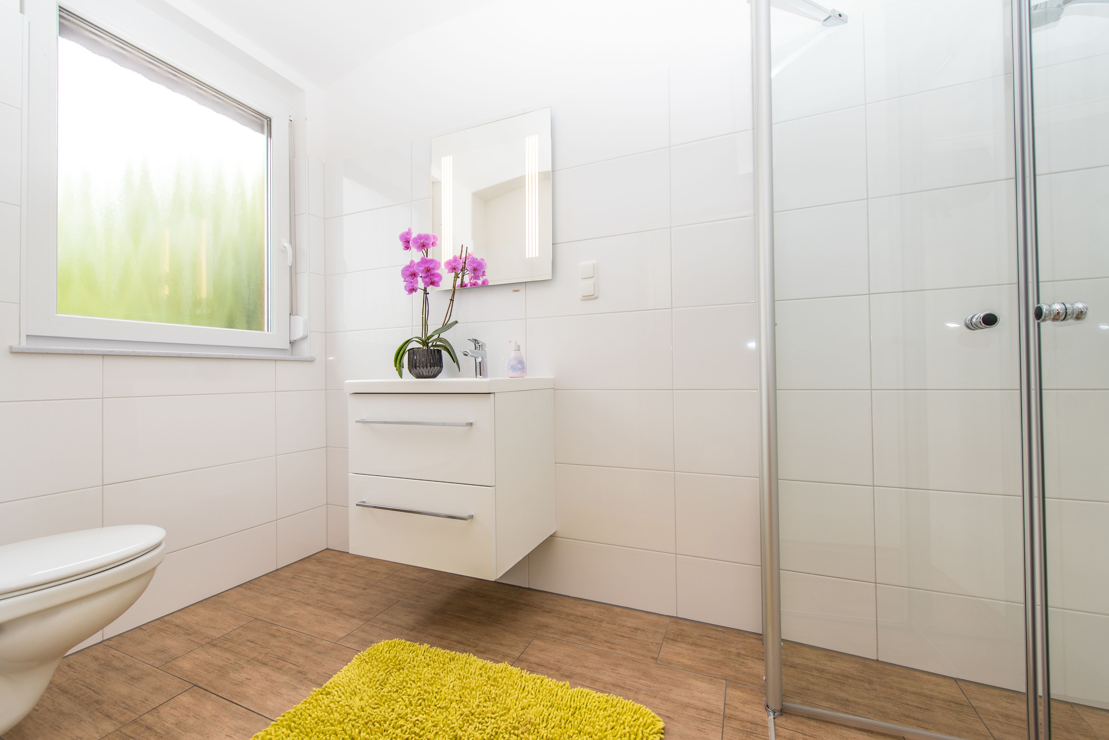 A modern bathroom with a toilet, a sink with a mirror, a shower, and a yellow rug on the floor.