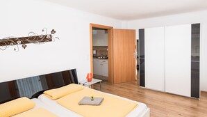 Et moderne hotelværelse med en stor seng, et lille bord med lampe, et garderobeskab og et tekøkken, der kan ses gennem en åben dør.