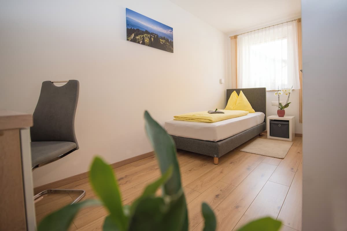Un dormitorio con cama, una silla, una mesita de noche, una ventana con cortinas y un cuadro en la pared.