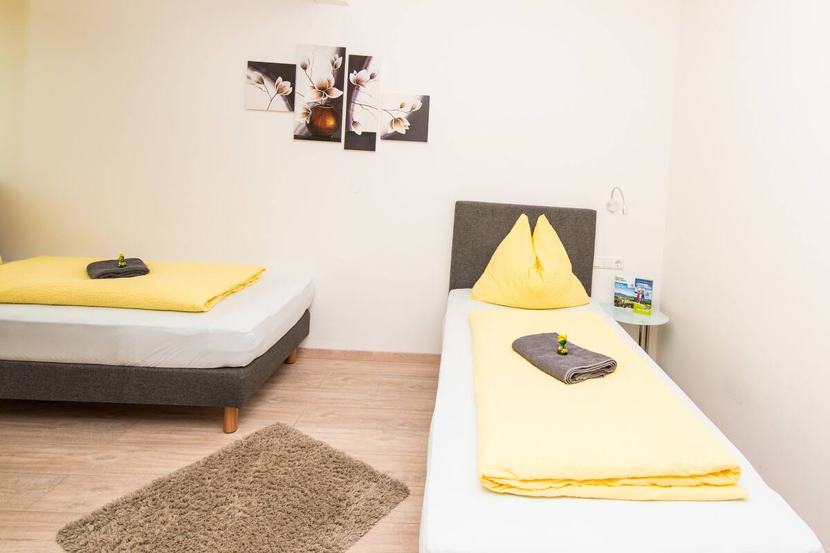 Un dormitorio con dos camas, cada una con una manta y almohada amarillas, una mesita con un libro y una lámpara en la pared.