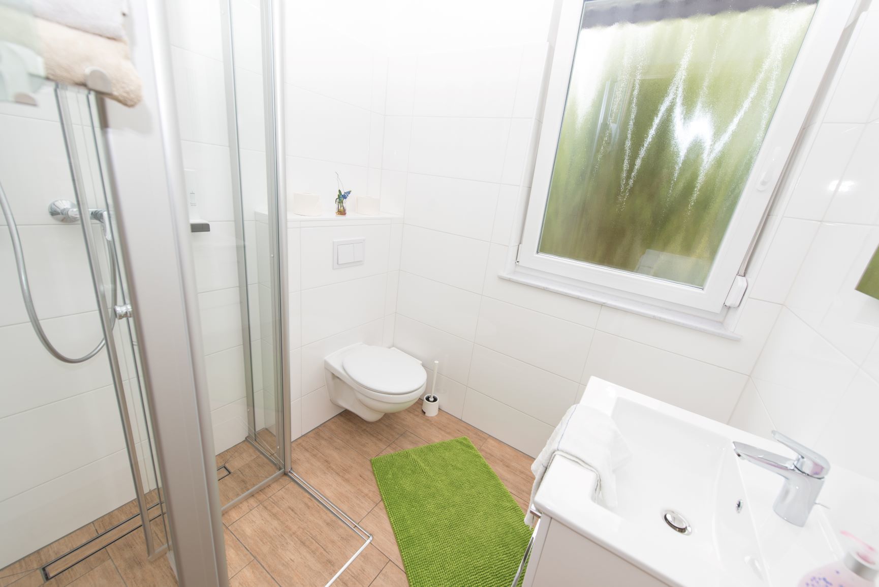 A bathroom with a glass shower enclosure, a toilet, a sink, and a window with a green curtain.