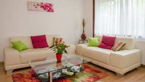 En stue med to lysebeige sofaer, farverige puder, et glas sofabord, en rød vase og et blomsterbillede på væggen.