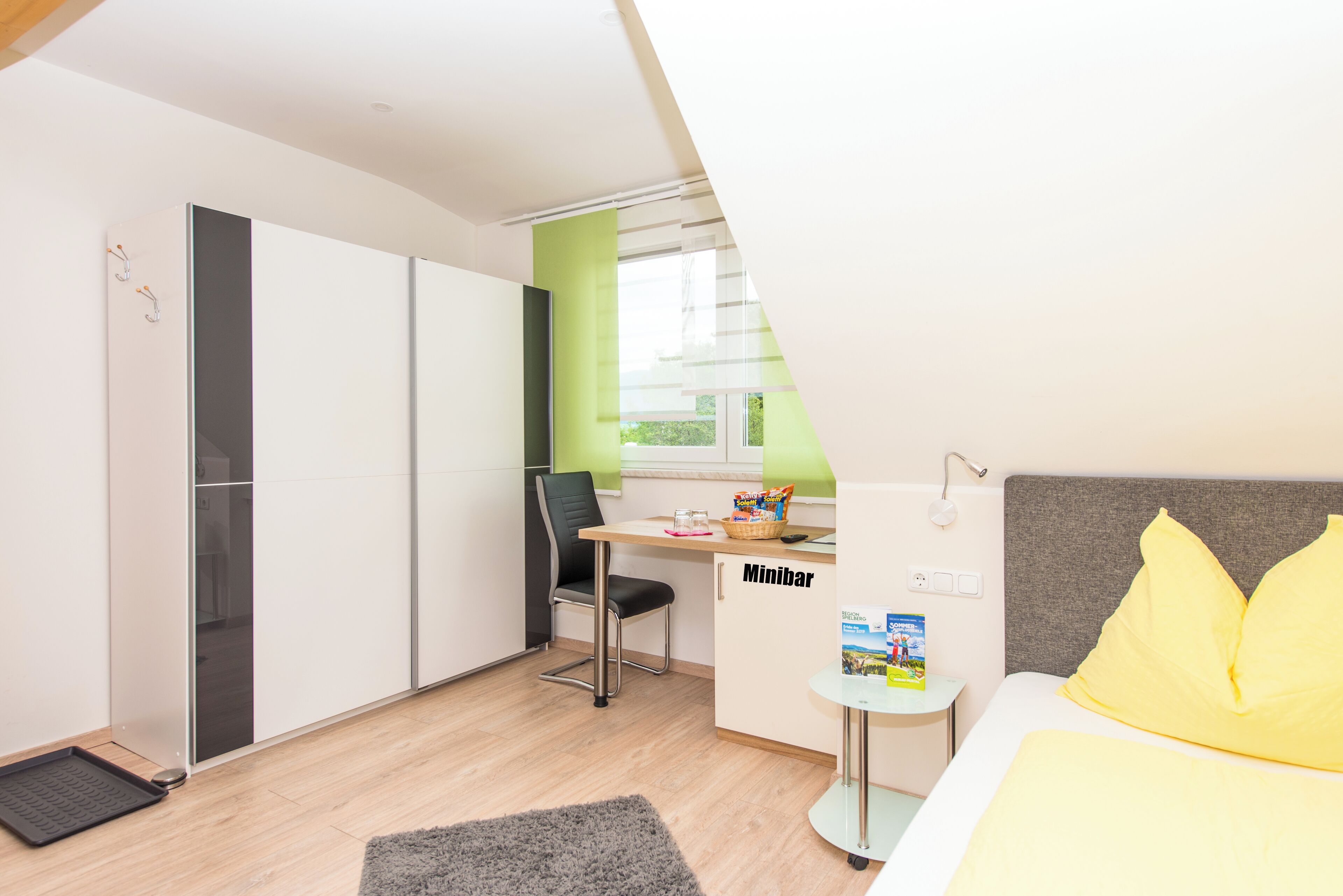 Habitación de hotel compacta con kitchenette, una cama con almohadas amarillas, una pequeña mesa con minibar y vistas a través de una ventana.