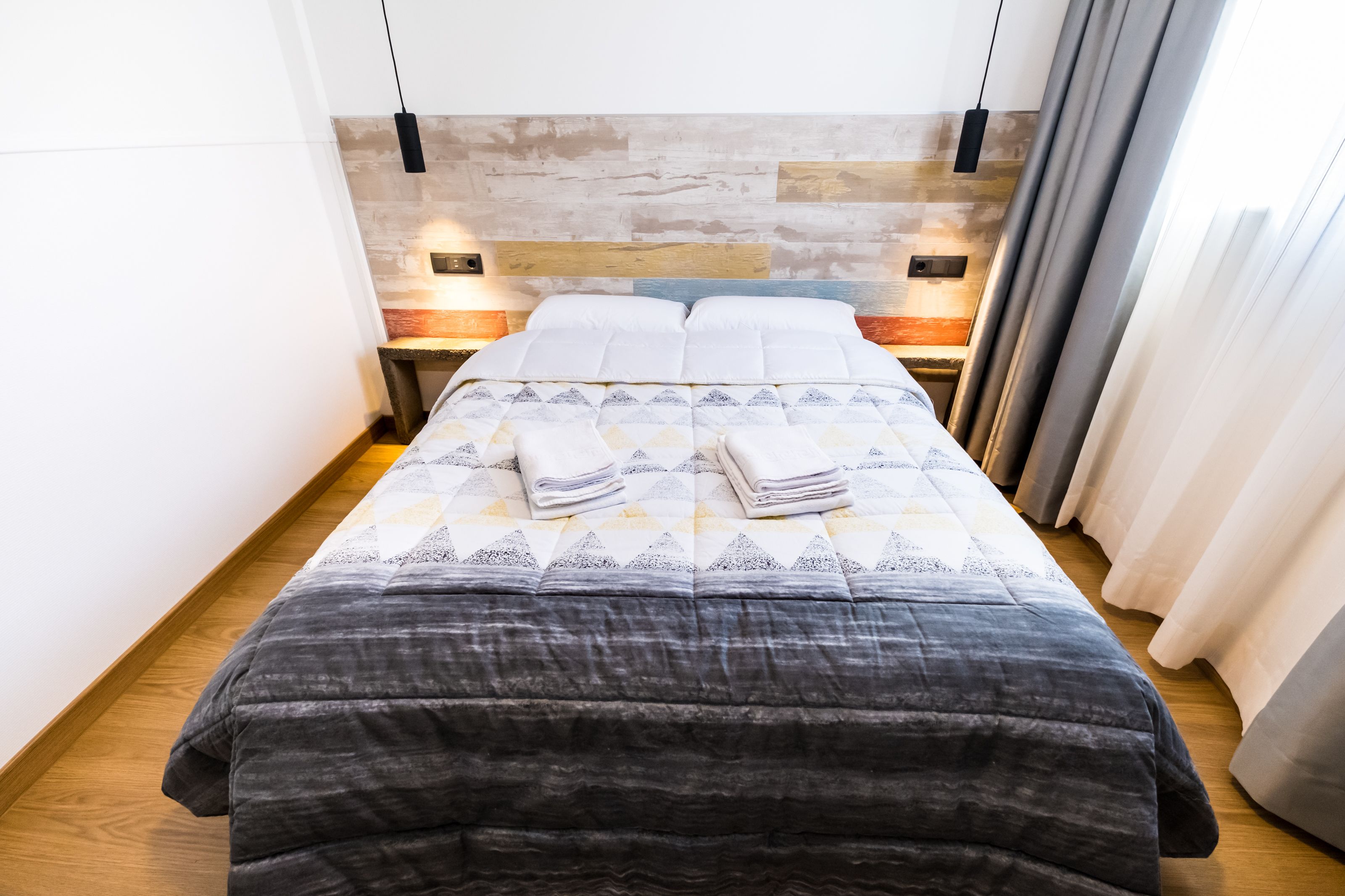 Ein ordentlich bezogenes Bett mit gemustertem Überwurf und Kissen, ein Holz-Kopfteil, Nachttischlampen und Blick ins Zimmerinnere.