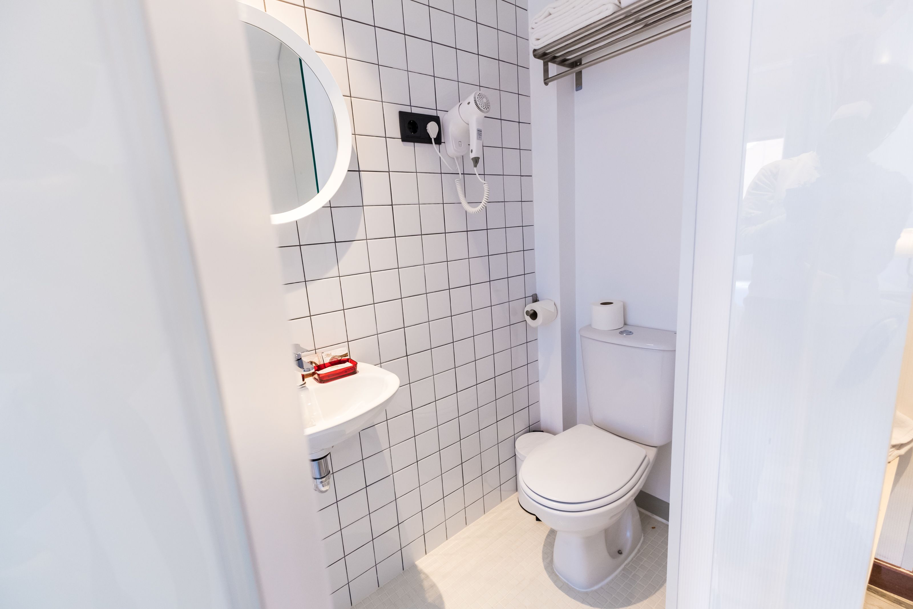 A modern bathroom with a white toilet, a sink with a red soap dispenser, and a hairdryer mounted on the wall.