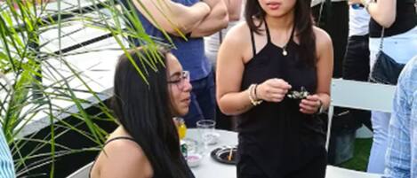 Un grupo de personas en un evento al aire libre con mesas y sillas, rodeado de vegetación y edificios al fondo.