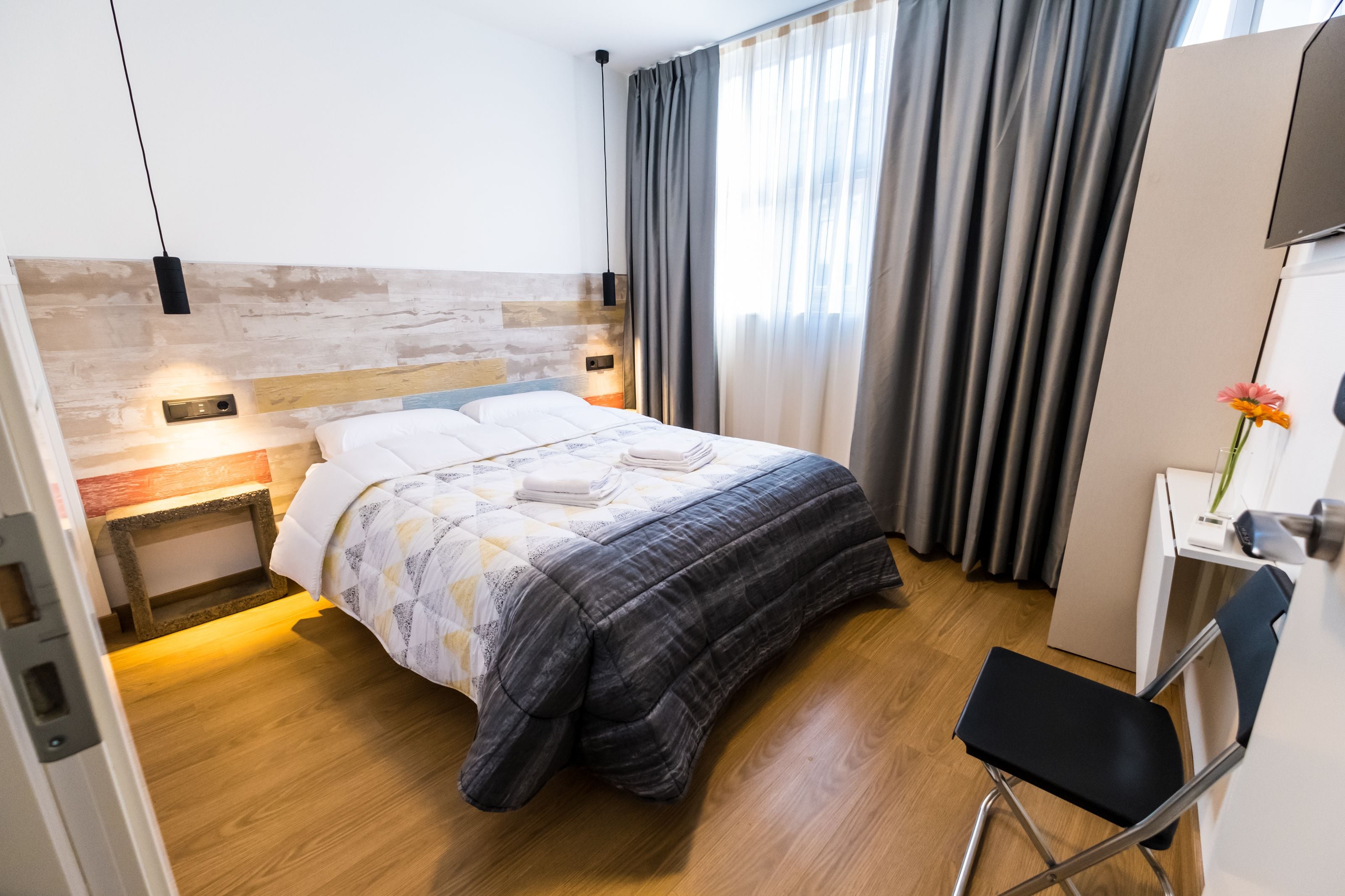 A modern hotel room with a wooden headboard, a bed with a quilt, a bedside table with a lamp, a desk with a chair, and a TV on the wall.