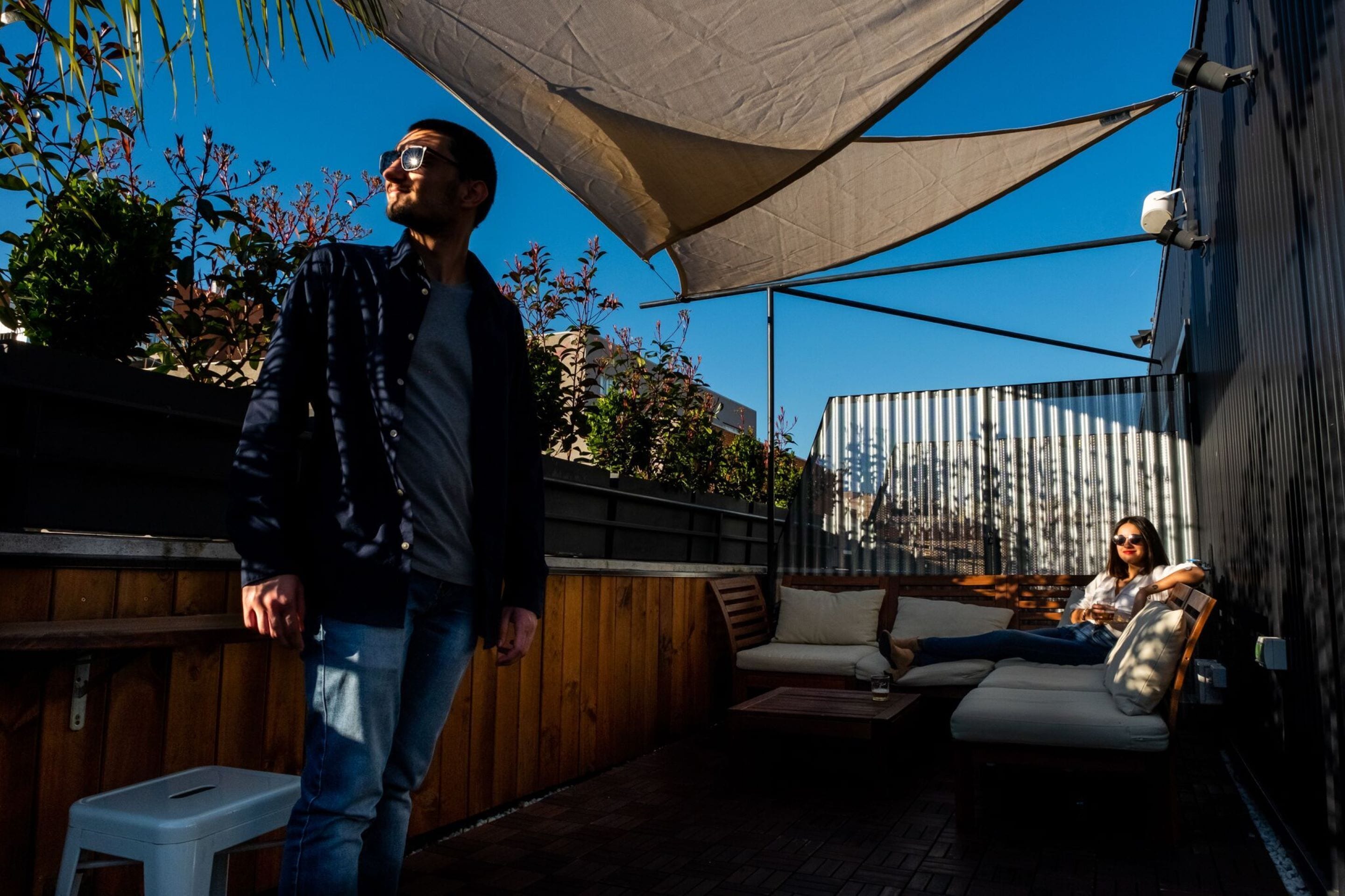 Un espace extérieur aménagé en terrasse, avec un sol en bois, un tabouret en plastique blanc, un coin salon confortable avec une table, et un grand auvent offrant de l’ombre.
