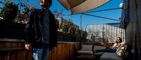 Un espace extérieur aménagé en terrasse, avec un sol en bois, un tabouret en plastique blanc, un coin salon confortable avec une table, et un grand auvent offrant de l’ombre.