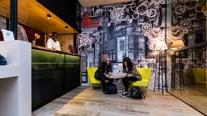 A reception area with a modern design, featuring a wooden reception desk, a yellow sofa, and a wall with mechanical gear patterns. - Far Home Bernabeu (Madrid)