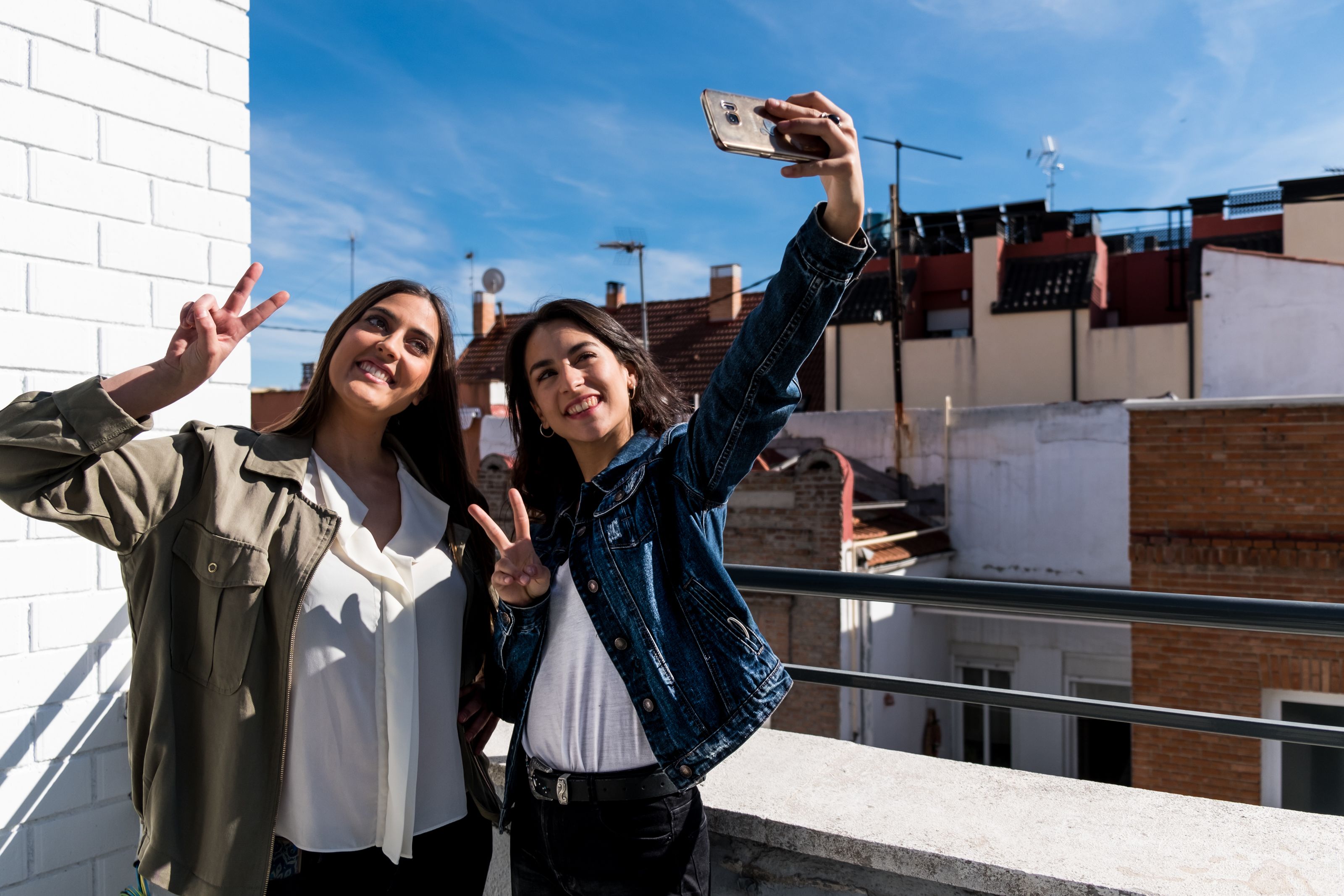 Two people taking a selfie on a balcony with a clear sky and buildings in the background.