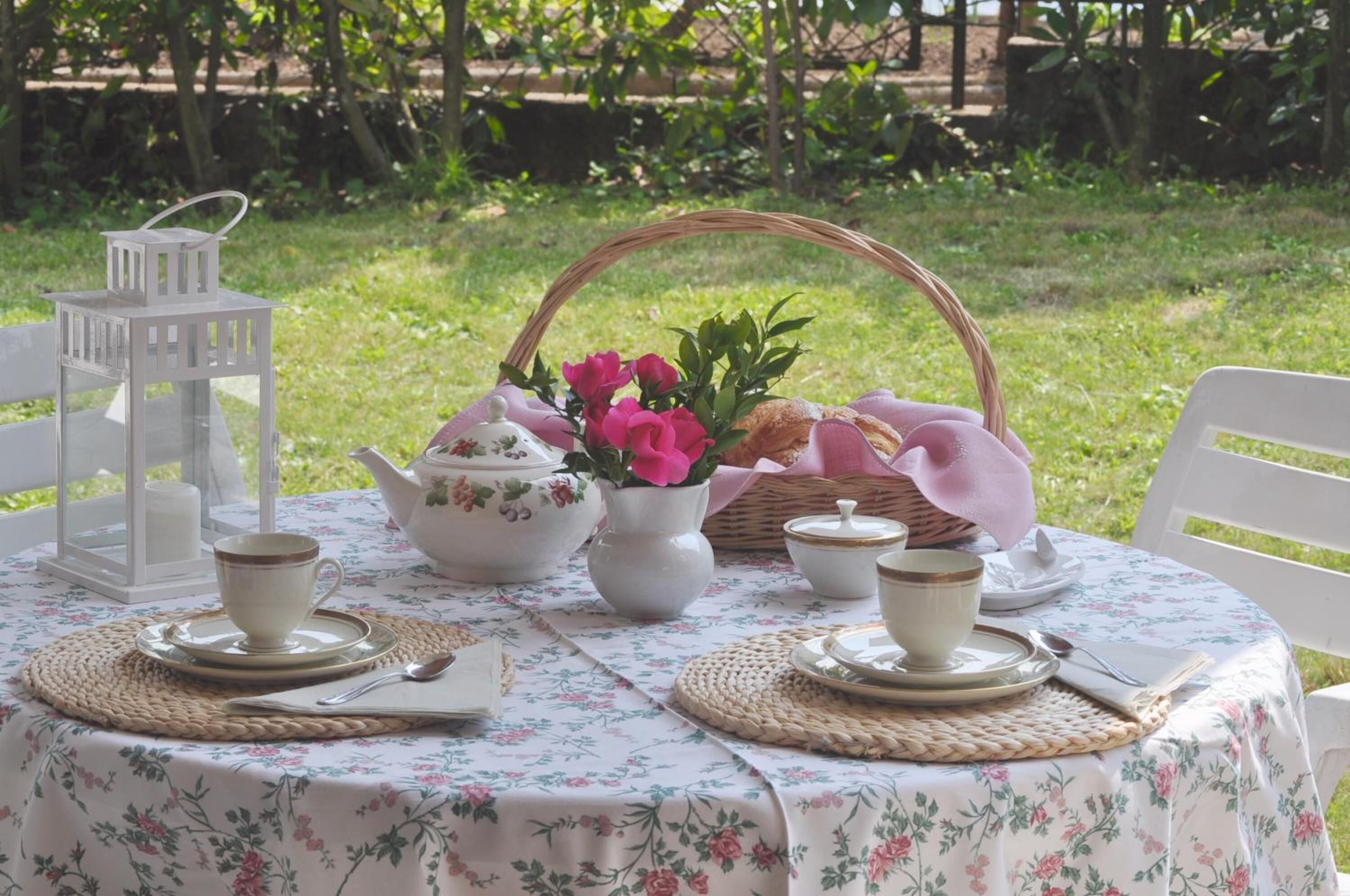 Una mesa colocada al aire libre con un mantel floral, tazas de té blancas, una tetera, una jarra, una cesta con pan y un farol.