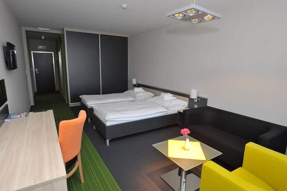 A hotel room with a bed, a desk, a chair, a small table with a vase, and a couch.