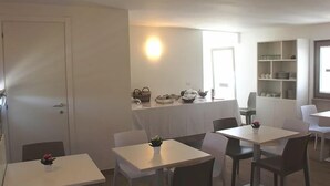 A dining area with white tables and chairs, a kitchen area with shelves, and a door. - I Cordari B&B (Vieste)