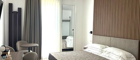A hotel room with a bed, a desk, a chair, a mirror, and a window with curtains.