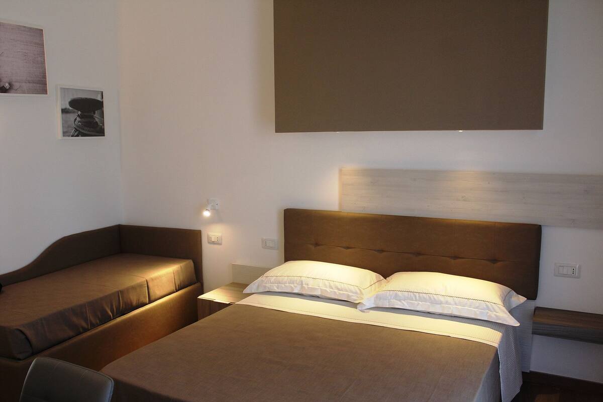 A hotel room with two beds, each with a headboard and pillows. There is a small table and a wall-mounted light.