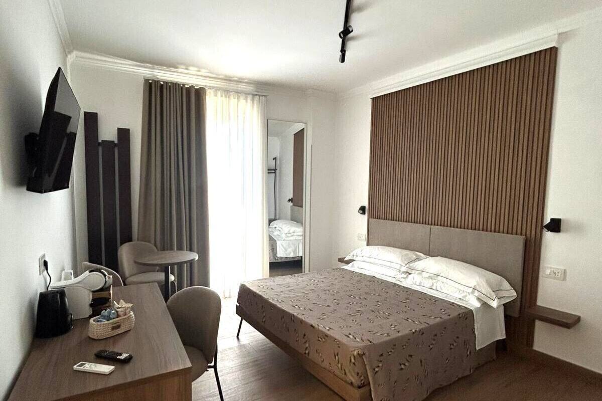 A hotel room with a bed, a desk, a chair, a TV mounted on the wall, and a window with curtains.