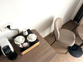 Una camera d'albergo con pavimento in legno, un tavolo nero con una macchina del caffè, un vassoio con tazze e una candela, e una sedia beige.