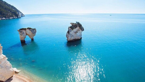 A coastal scene with two prominent rock formations in clear blue water, surrounded by a sandy beach and a distant mountain. - I Cordari B&B (Vieste)