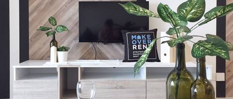A modern living room with a herringbone wood wall, a flat-screen TV, a shelf with decorative items, and a tray with a glass, candles, and a plant.