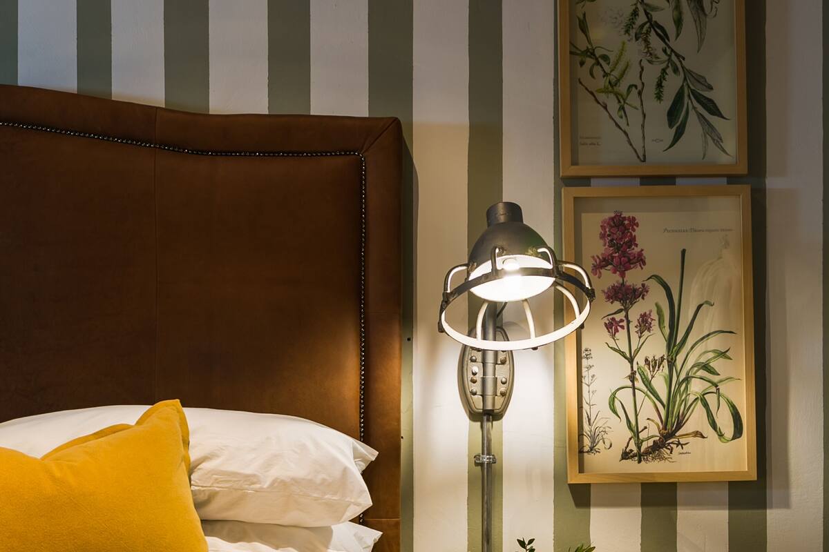 Una cama bien hecha con cabecera marrón, almohada amarilla y colcha verde. Hay una mesita de noche con una lámpara e impresión botánica enmarcada en la pared.