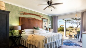Een slaapkamer met een groot bed, een plafondventilator, een nachtkastje met een lamp en uitzicht op het omliggende landschap.