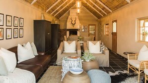 Een ruime woonkamer met een hoge plafond, een open haard en diverse zithoeken, waaronder een bank, stoelen en een ronde tafel.