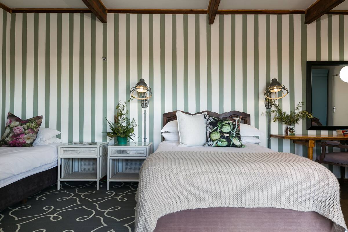 Habitación de hotel con papel tapiz a rayas, una cama grande con una manta blanca de punto, dos mesitas de noche con lámparas y un escritorio pequeño con una silla.