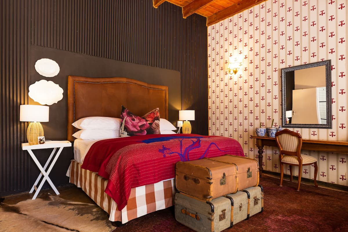 Un dormitorio con una cama grande, dos mesitas de noche con lámparas, un cabecero de madera, un escritorio con silla y papel tapiz estampado.
