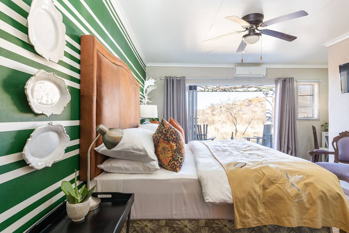 Habitación de hotel con una cama, ventilador de techo, ventana con cortinas y vistas a árboles.
