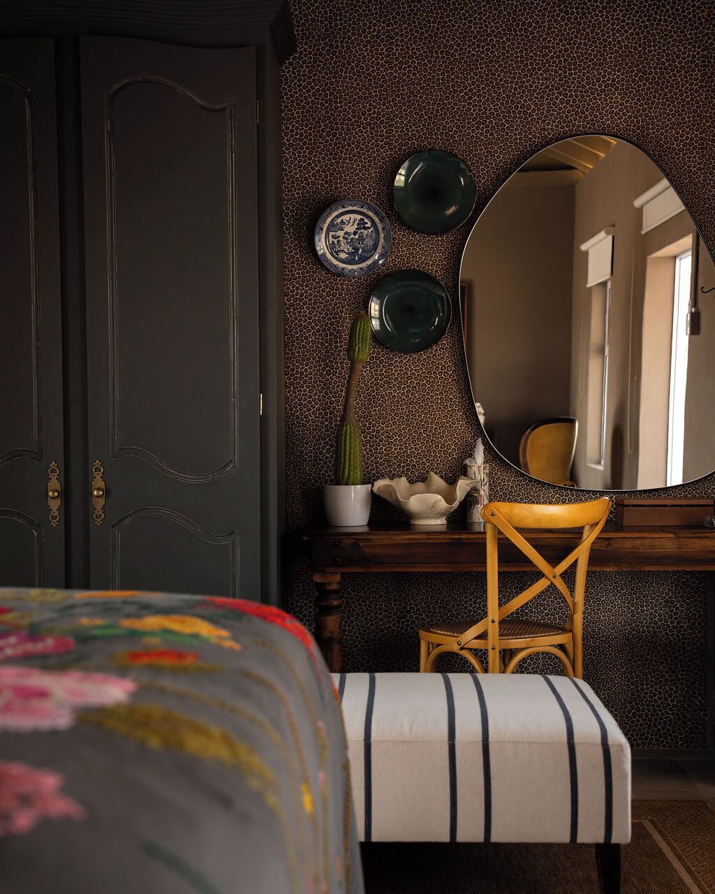 A room with a dark door, a round mirror, a wooden chair, a small table with a potted plant, and a striped bench.