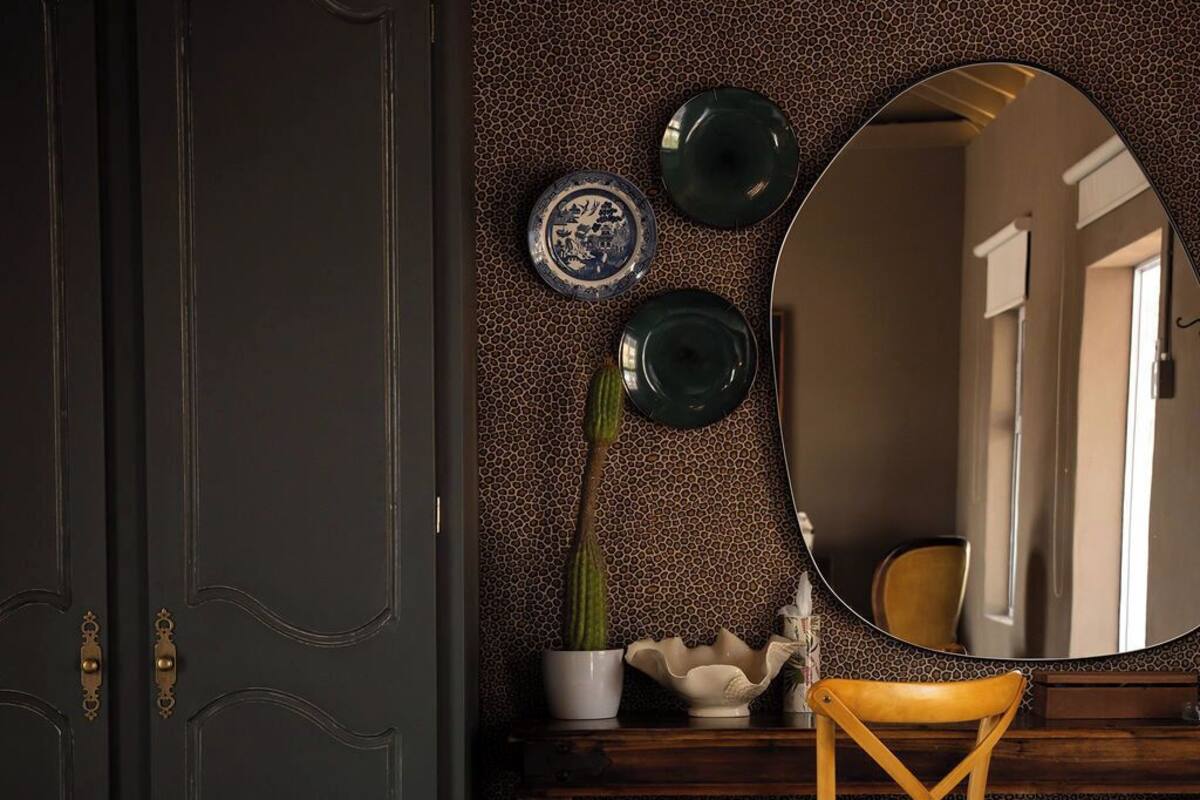 Una habitación con una puerta oscura, un espejo redondo, una silla de madera, una mesita con una planta en maceta y un banco a rayas.