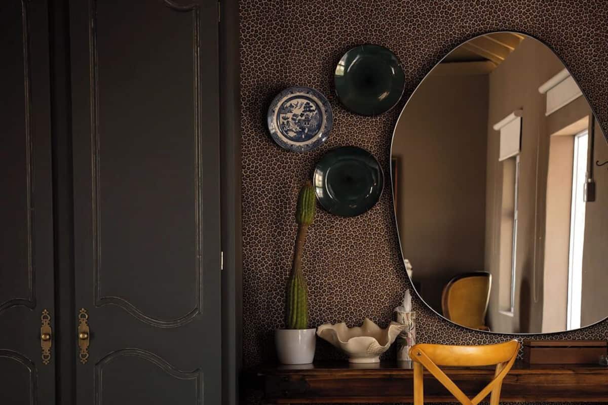 A room with a dark door, a round mirror, a wooden chair, a small table with a potted plant, and a striped bench.